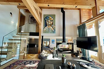 Bright, stylish open-plan living space with exposed wooden beams, a sleek glass staircase, modern suspended fireplace, and a large abstract portrait painting.