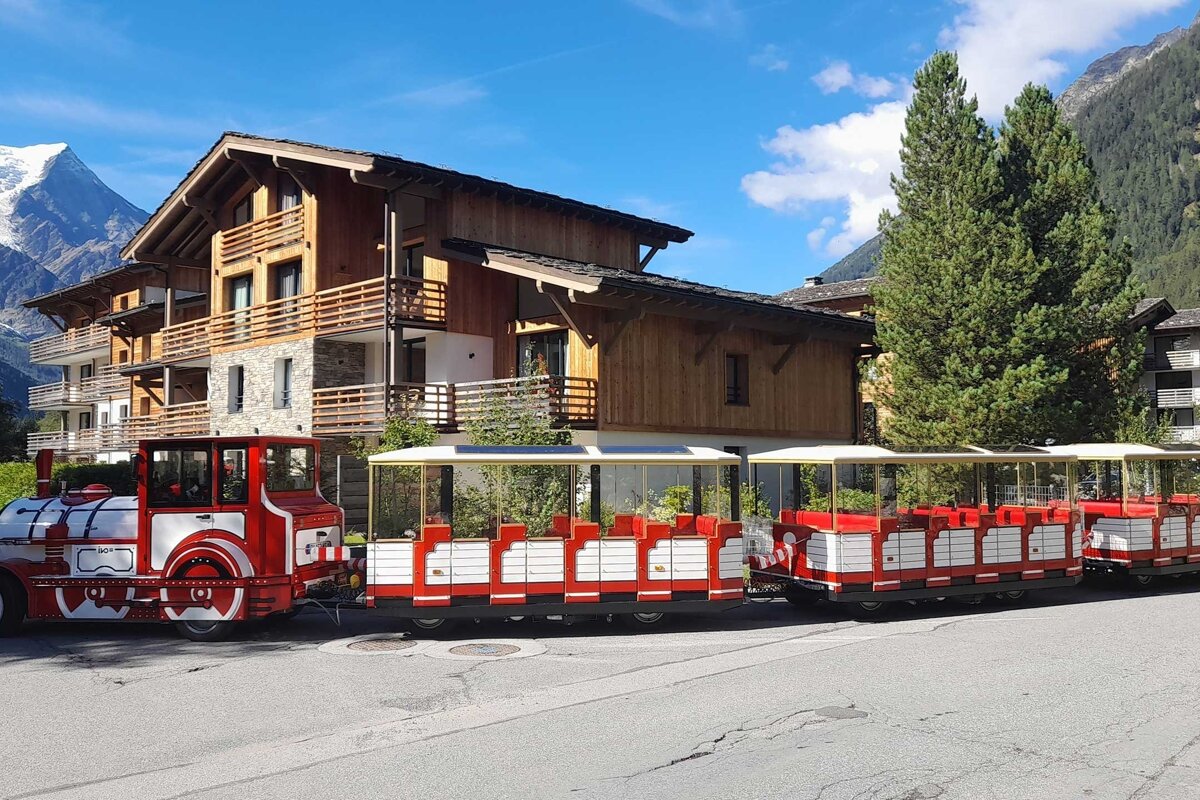 A red and white tourist train is parked on a street in front of Alpine chalets and snow-capped mountains under a clear blue sky.