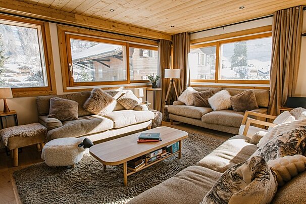 A cozy, sunlit chalet living room with wood paneling, plush sofas, and large windows framing a snowy mountain village. A sheep ottoman adds charm.