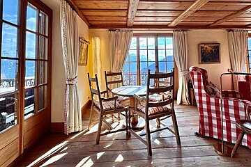 Cozy chalet interior with wood ceilings, a wooden dining set, red checkered armchair, and large windows offering scenic mountain views.