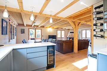 An inviting open-plan chalet interior with a sleek kitchen island, dining table, and a living room featuring a fireplace, brown leather sofas, and exposed wooden beams.