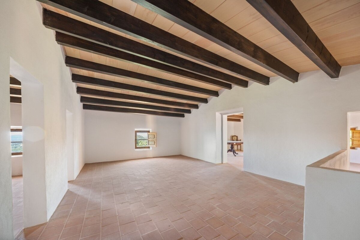 An empty room with white walls, terra cotta floor, and dark exposed wooden ceiling beams. Small windows offer glimpses outside.