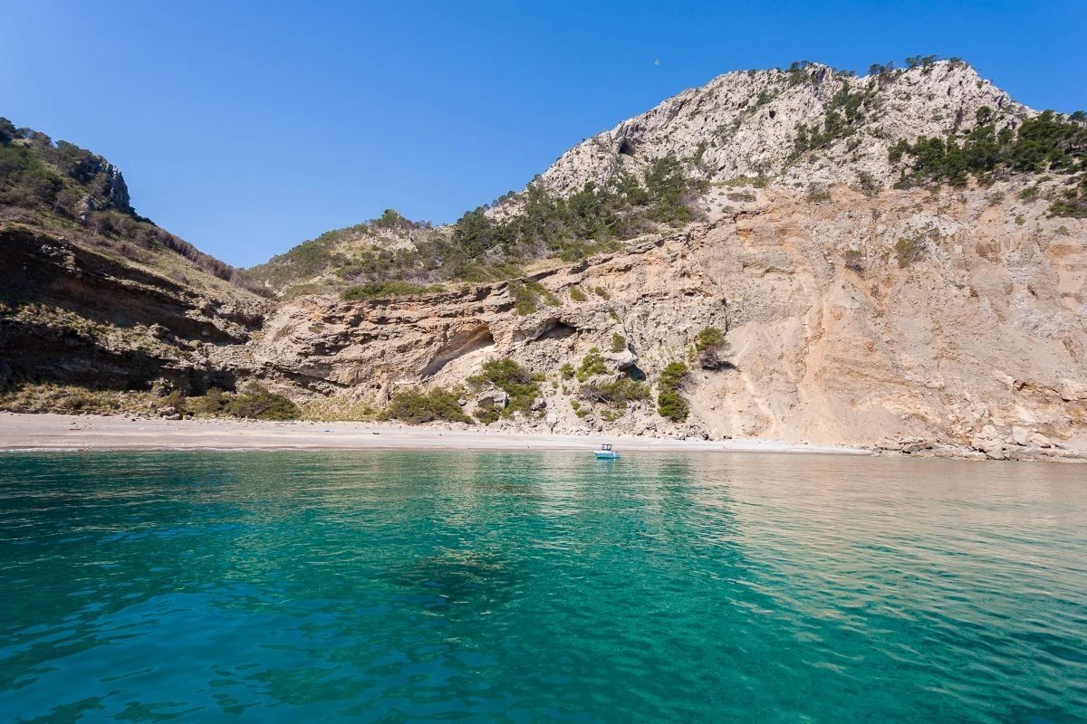 Azure sea meets a sandy beach at the base of towering, tree-covered cliffs under a clear blue sky. A small boat floats by the shore.