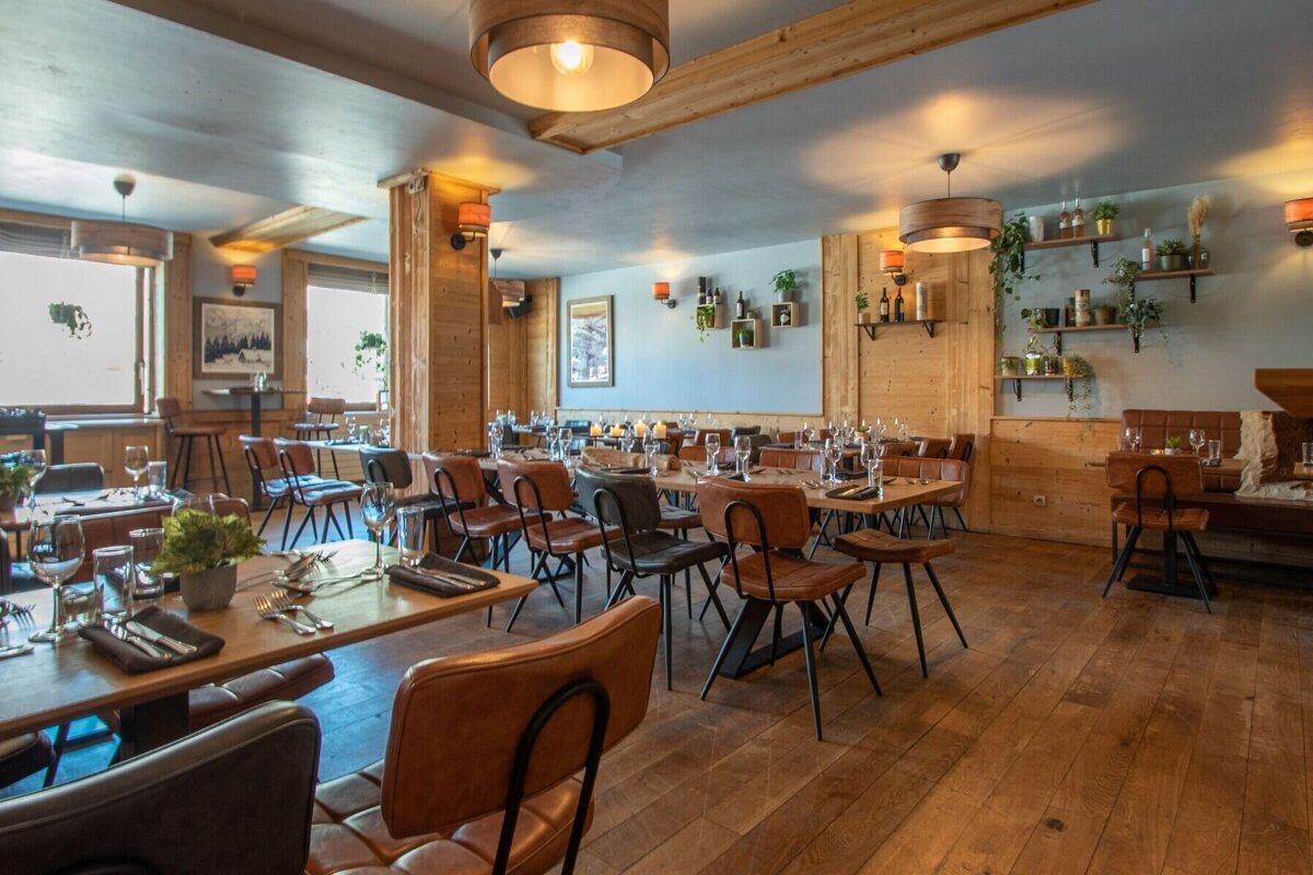 A warm, rustic restaurant interior with wooden walls, floors, and set tables with brown chairs. Pendant lights and wall sconces create a cozy ambiance.