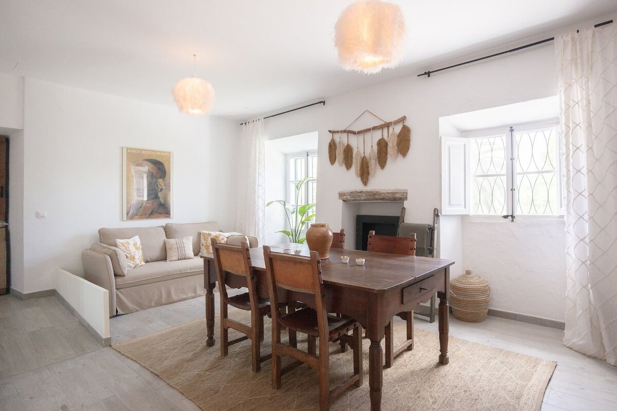 A bright, rustic living/dining room with a wooden table, cozy sofa, fireplace, and fluffy pendant lights in a white-walled space.