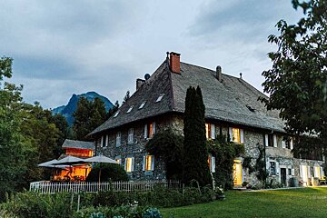 A charming stone house with illuminated windows at dusk, set against mountains, surrounded by lush greenery and a cozy outdoor patio.