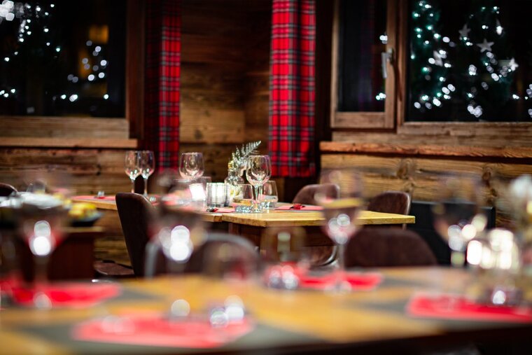 A cozy, festive restaurant interior with wooden walls, red plaid curtains, set tables, and blurry Christmas lights outside the windows.