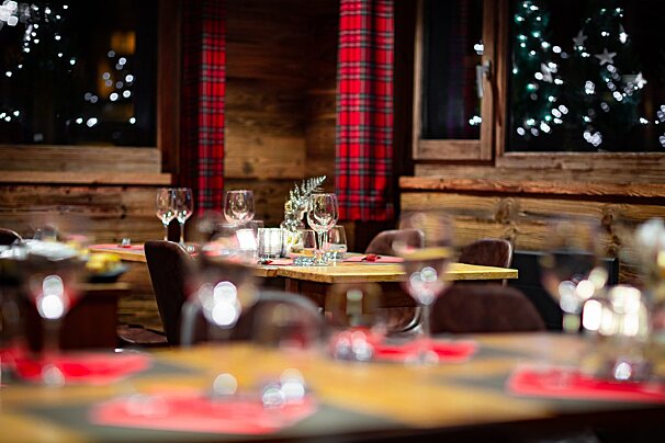 A cozy, festive restaurant interior with wooden walls, red plaid curtains, set tables, and blurry Christmas lights outside the windows.