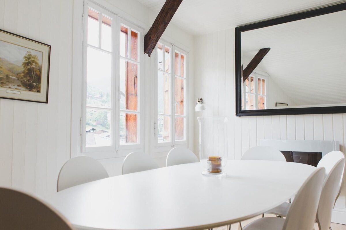 A bright, white minimalist dining room features an oval table, modern chairs, large windows, an exposed wooden beam, and a big wall mirror.