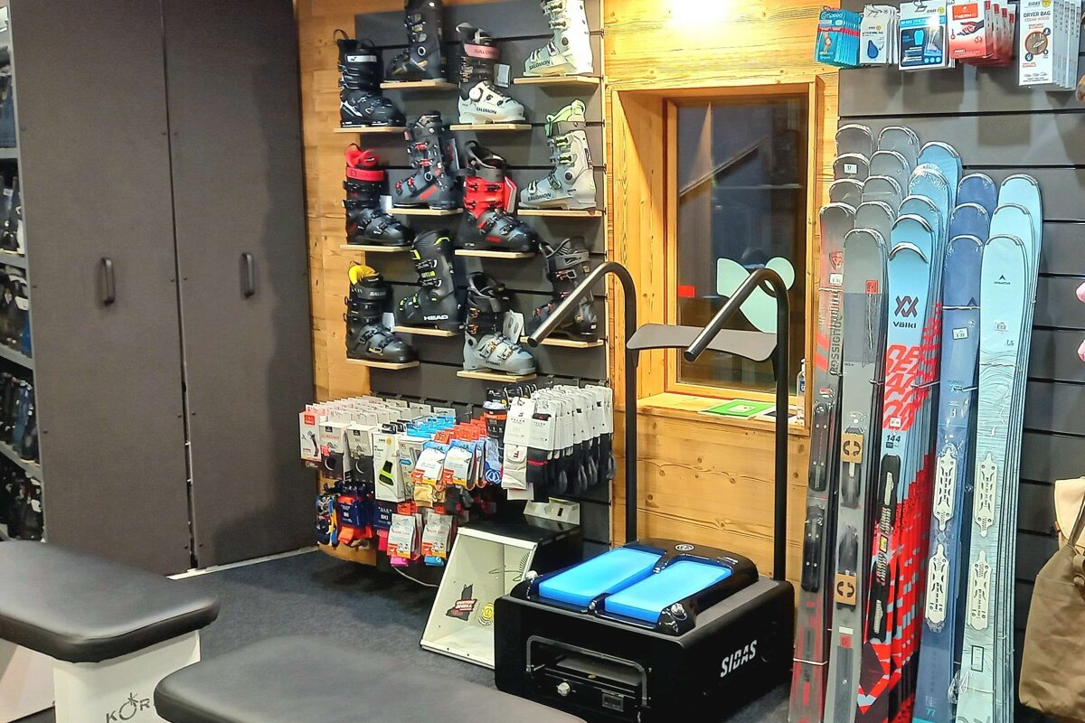 A ski shop displays shelves of ski boots, rows of skis, ski accessories, and a SIDAS boot fitting machine in a rustic setting.