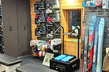 A ski shop displays shelves of ski boots, rows of skis, ski accessories, and a SIDAS boot fitting machine in a rustic setting.