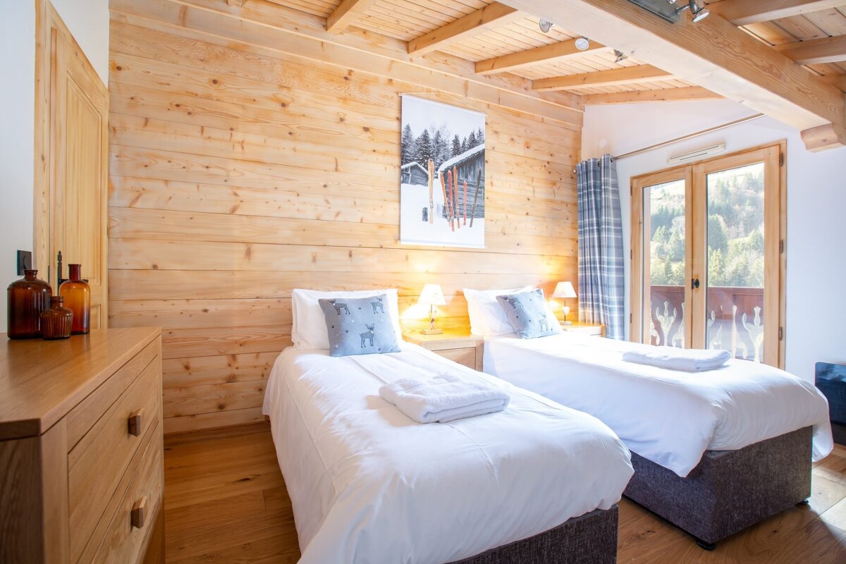 A cozy twin bedroom with light wood paneling, ski art, and deer pillows. A balcony offers a forest view, creating a warm, alpine chalet feel.