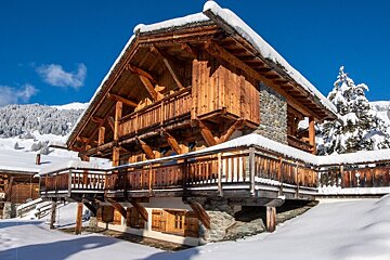 A snow-covered wooden chalet with balconies under a bright blue sky, set in a sunny winter mountain landscape.