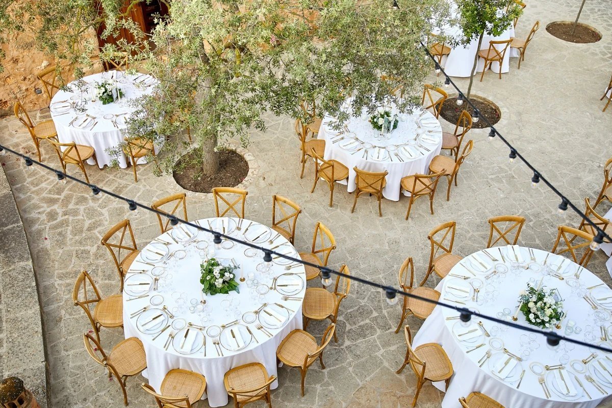 Elegant outdoor event setup: round tables with white linens, wooden chairs, lush trees, and festive string lights in a stone courtyard.