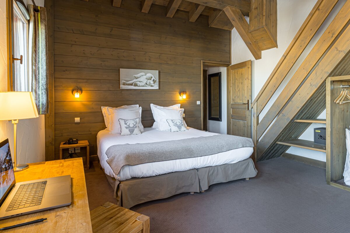 A cozy, rustic bedroom with wood-paneled walls, exposed beams, a large bed, and a laptop on a wooden desk. Warm lighting enhances the inviting atmosphere.