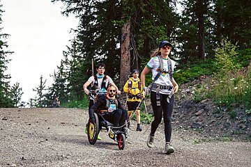 Four people on a forest trail, three assisting a person in a specialized wheelchair, moving uphill during an outdoor event.