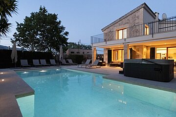 An inviting stone villa at dusk, featuring a sparkling lit pool, hot tub, and comfortable lounge chairs, creating a serene outdoor oasis.