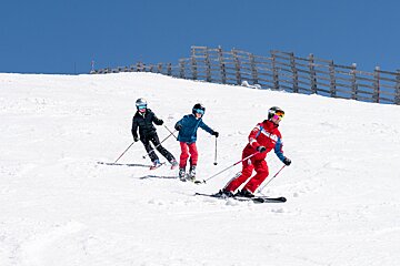 Three people ski down a bright white mountain slope under a clear blue sky, with a wooden fence visible in the background.