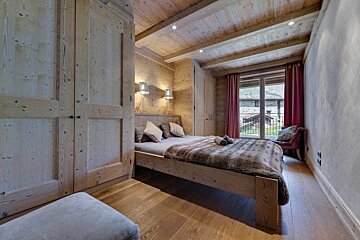 A cozy, chalet-style bedroom with wooden walls, ceiling, floor, and wardrobe. Features a bed with a furry throw and a window with red curtains.
