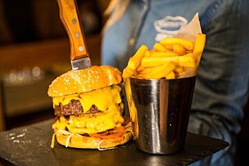 A huge double cheeseburger with a knife in it, served with a metal cup overflowing with fries, presented on a dark platter.