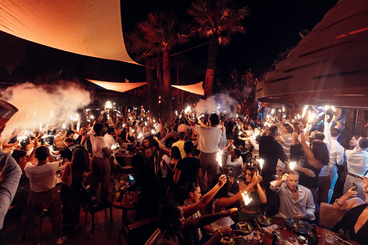 A lively, crowded outdoor night party with many people holding sparklers and creating a smoky, festive atmosphere under palm trees.