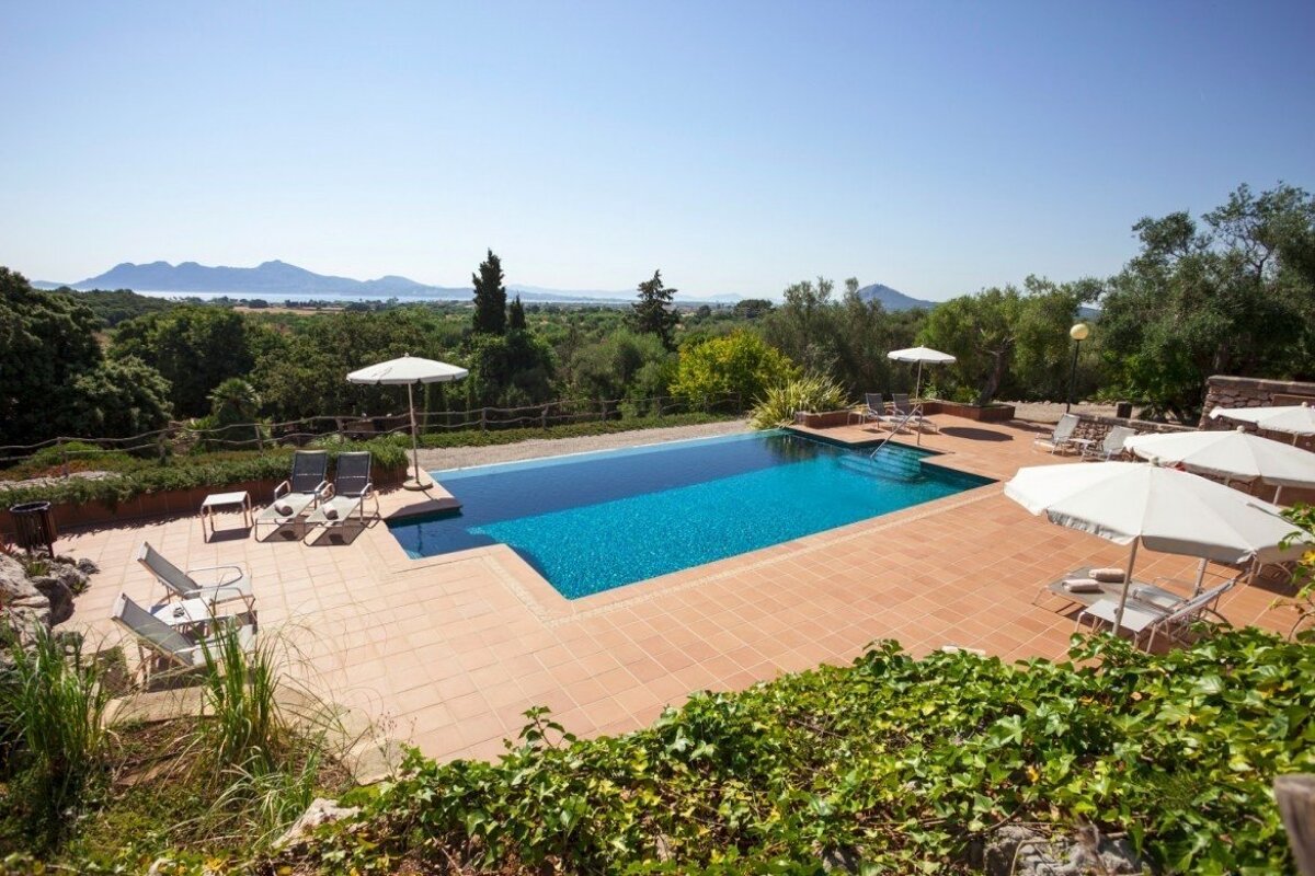 An inviting infinity pool with a terracotta deck, lounge chairs, and umbrellas overlooks lush greenery, distant mountains, and a clear blue sky.