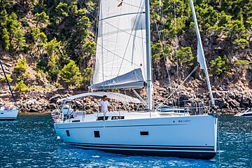 A sleek white sailboat with its sails unfurled cruises on clear blue waters alongside a lush green, rocky coast with two people aboard.