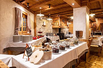 A cozy, rustic breakfast buffet room with wooden beams and stone walls. Tables are laden with cereals, dried fruits, drinks, and hot food items.