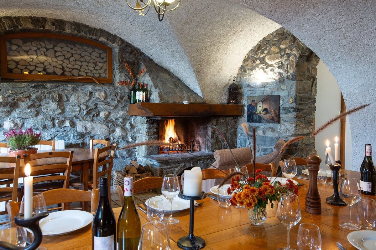 Cozy, rustic dining room with stone walls, vaulted ceiling, and a roaring fireplace. A wooden table is set with wine, candles, and flowers.