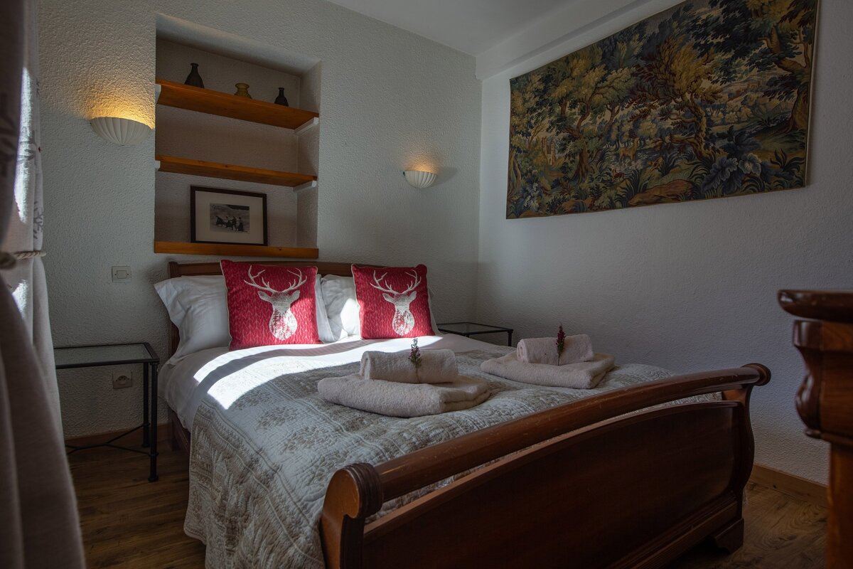 A cozy bedroom features a wooden bed with red deer pillows, white walls, a forest tapestry, and recessed shelves. Sunlight streams across the bed.