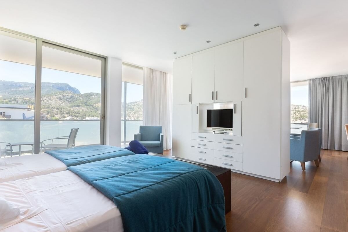 Modern hotel room with twin beds, large window overlooking mountains & water, white wardrobe with built-in TV, blue armchair, and wooden floor.