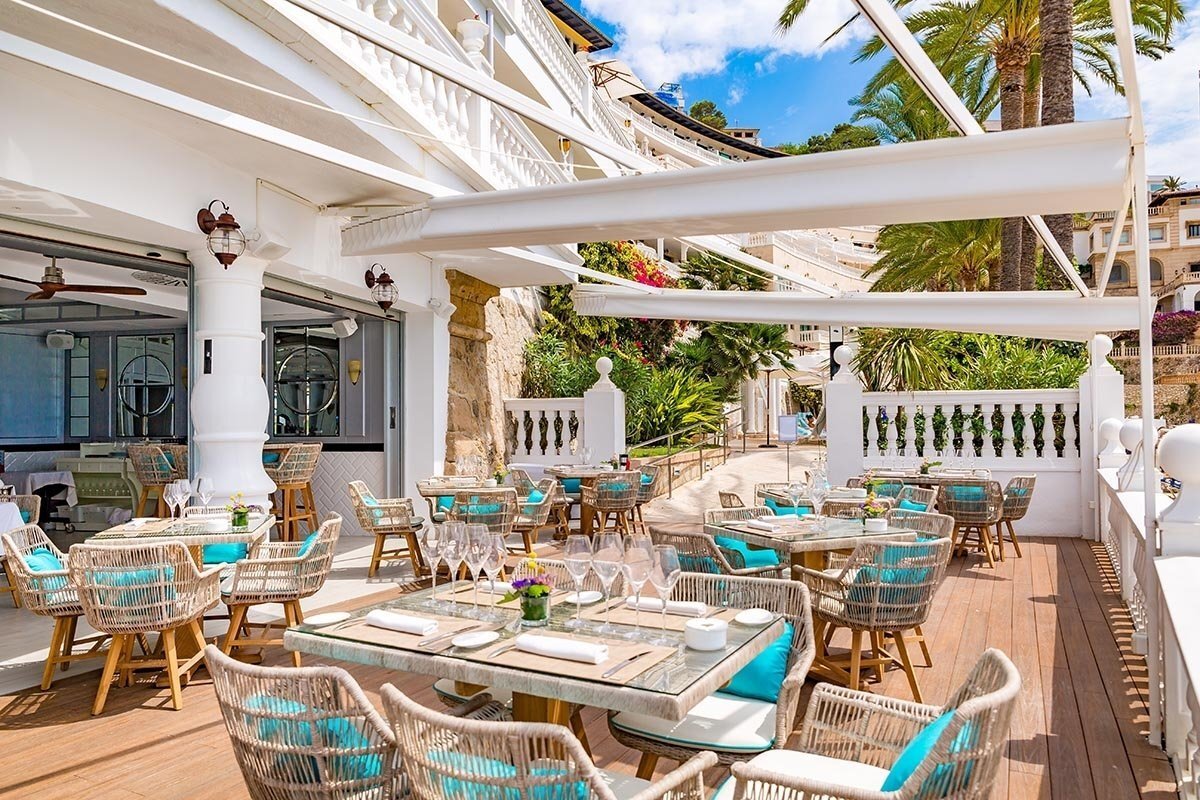 Chic outdoor restaurant patio with wicker chairs & blue cushions under white awnings, surrounded by a resort building, palm trees, and lush greenery.