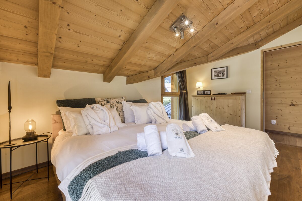 A warm, rustic bedroom with a slanted wooden ceiling and exposed beams. A large bed features white linens, bathrobes, and rolled towels, creating a cozy chalet feel.