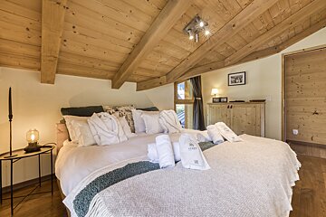 A warm, rustic bedroom with a slanted wooden ceiling and exposed beams. A large bed features white linens, bathrobes, and rolled towels, creating a cozy chalet feel.