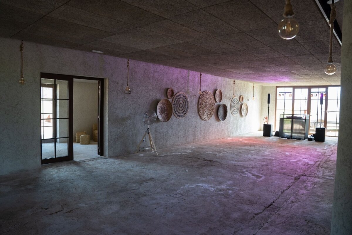 An empty, industrial-style hall with concrete walls, woven circular decor, a DJ setup, and purple ambient lighting. Glass doors lead to other rooms.