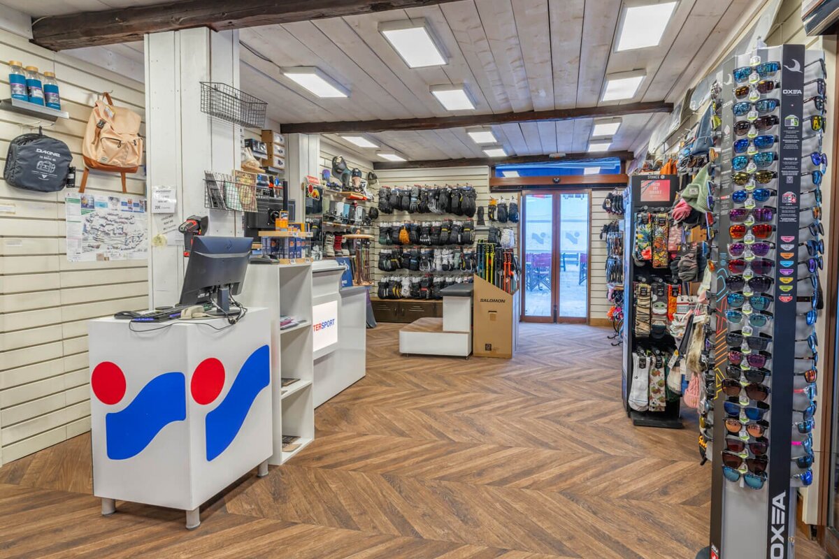 Brightly lit sports equipment shop interior with displays of sunglasses, gloves, and outdoor gear. A reception desk and wooden herringbone floor are visible.