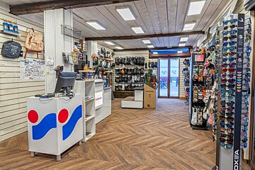 Brightly lit sports equipment shop interior with displays of sunglasses, gloves, and outdoor gear. A reception desk and wooden herringbone floor are visible.
