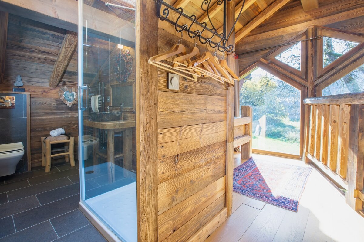 A rustic, woody interior featuring a modern glass shower, toilet, and hangers, with large windows overlooking the outdoors in a chalet-style setting.