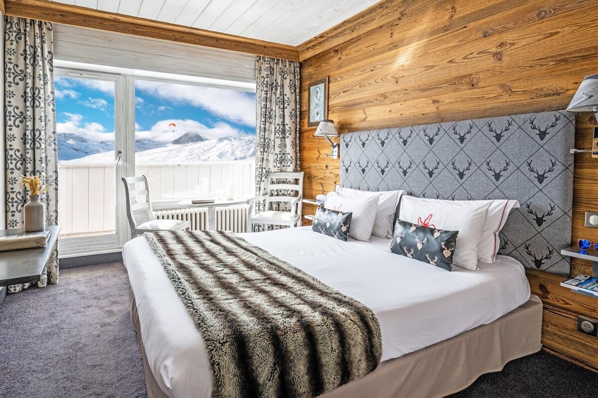 A cozy, wood-paneled ski chalet bedroom with two beds, fur throws, and a large window overlooking snowy mountains and buildings. A deer head adorns the wall.