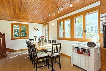 A cozy dining room with a large wooden table, black chairs, and a wooden paneled ceiling. Large windows reveal a lush outdoor view, while ski decor adds a rustic touch.