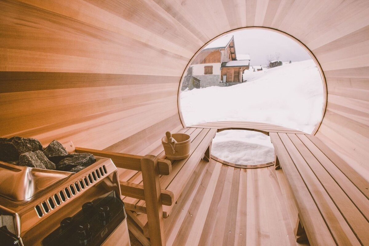 Inside a wooden barrel sauna, a circular window reveals a snowy mountain landscape with a distant house. A heater, benches, and bucket are visible.