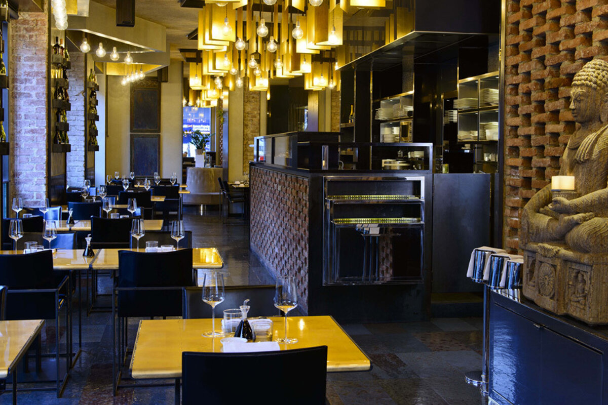 A stylish, dimly lit restaurant interior. Features modern tables, brick walls, unique suspended lighting, and a serene Buddha statue, creating an Asian-inspired ambiance.