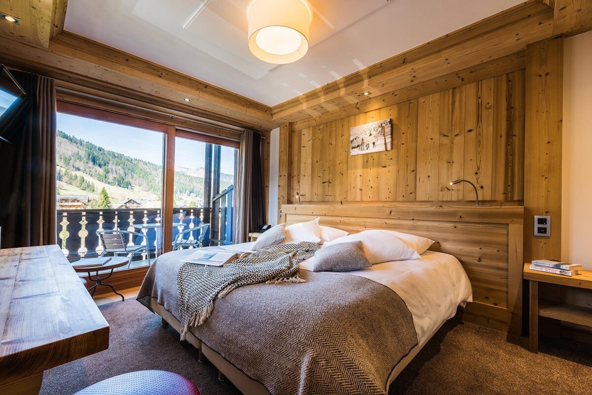 A cozy, wood-paneled bedroom features a large bed, warm lighting, and a private balcony offering stunning views of a lush mountain valley.