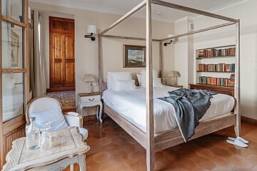 A cozy, rustic bedroom with a wooden four-poster bed, white bedding, terracotta tile floor, built-in bookshelf, and an armchair by French doors.