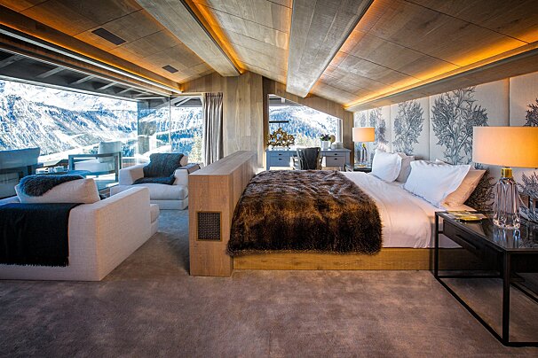 Luxurious bedroom with a large window overlooking snowy mountains, featuring a modern design, fur throw, and integrated sitting area.