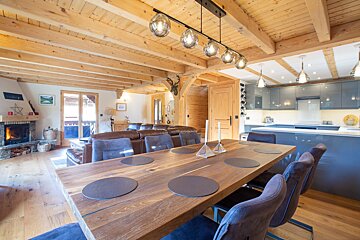 A spacious open-plan chalet interior with a large wooden dining table, a cozy living room fireplace, and a modern grey kitchen under exposed wooden beams.