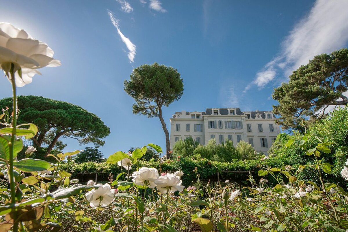 A large white building is surrounded by flowers and trees