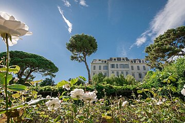 A large white building is surrounded by flowers and trees