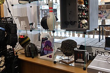 A retail display case showcases winter sports apparel including gloves, beanies, helmets, and sunglasses, with a snowy outdoor view in the background.