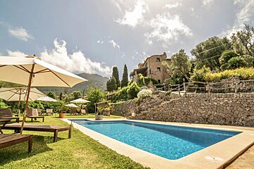 A sunny stone villa with a bright blue swimming pool, surrounded by lush gardens, stone walls, umbrellas, and distant mountains.